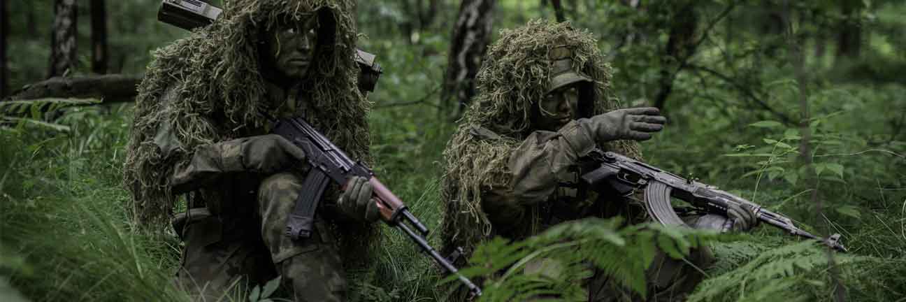 Two airsoft players in camouflage gear with airsoft rifles in a forest setting